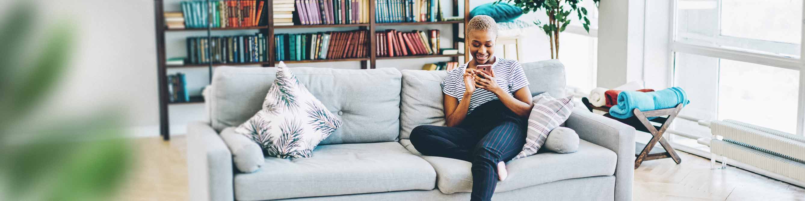 Frau sitzt mit Handy in der Hand auf einer Couch vor bunter Bücherwand.